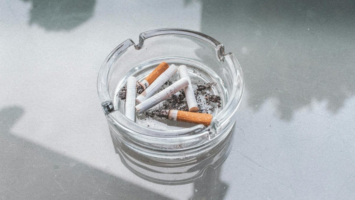 A close-up view of used cigarette butts in a glass ashtray on a reflective surface, highlighting indoor smoking.