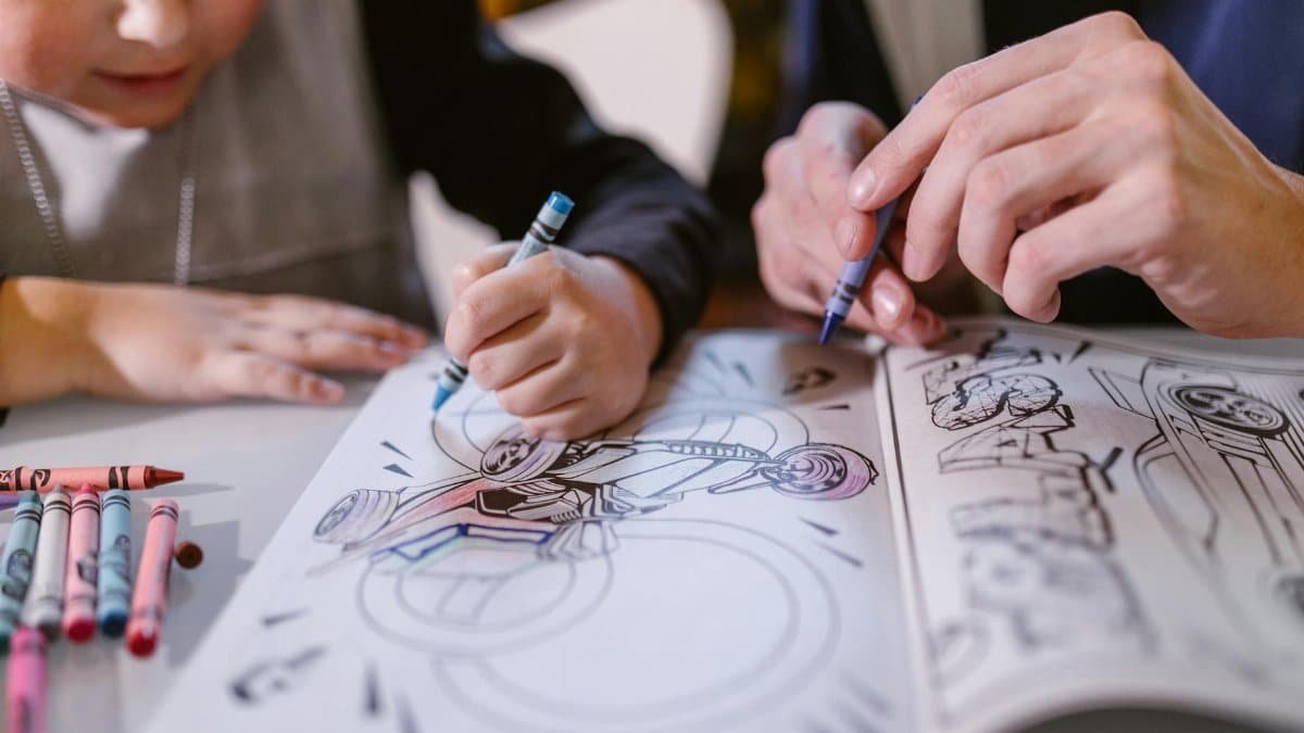 Child and an adult coloring a biker image in a coloring book, using crayons.