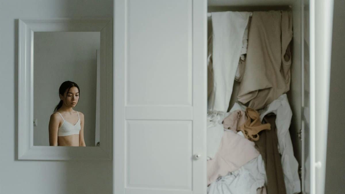 A young woman reflects in the mirror beside an unorganized closet.