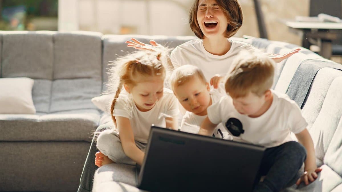 Stressed woman asking for help while sitting with small children playing on laptop together on sofa in living room