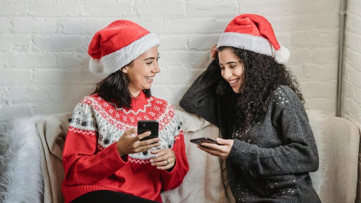 Cheerful young Hispanic female friends in warm jumpers and Santa hats sitting on small sofa and browsing smartphones while spending Christmas holiday together