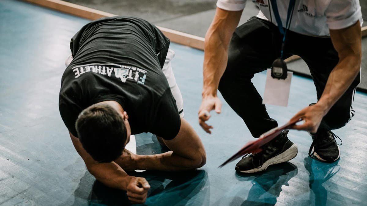 An athlete being assisted by a coach during an indoor training session on a blue floor.