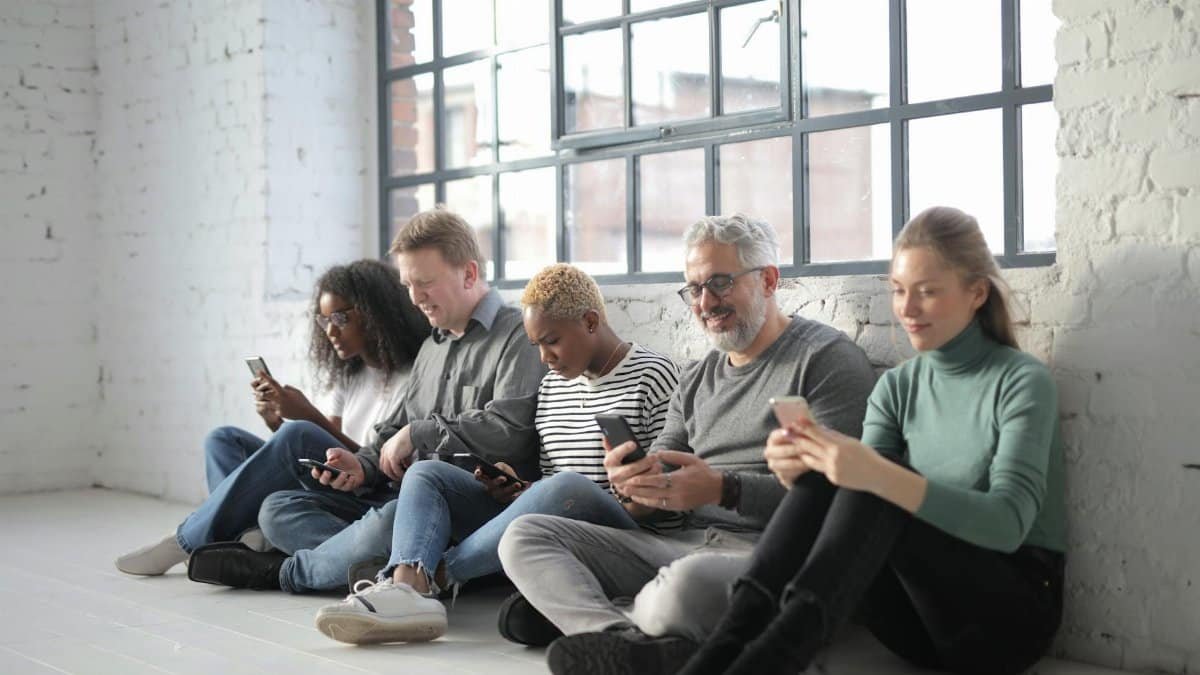 Smiling coworkers of different ages in casual outfit browsing internet on cellphones while sitting with crossed legs near brick wall and fenced window during daytime