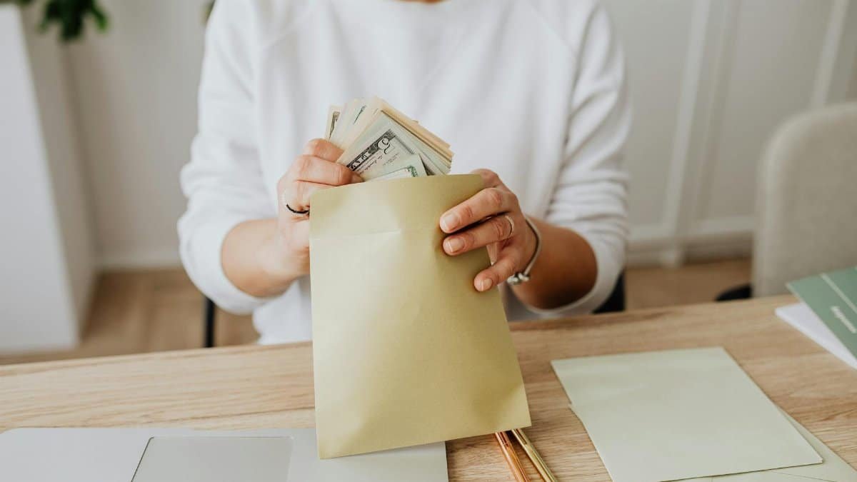 Close-up of person holding envelopes with cash at a wooden desk indoors.