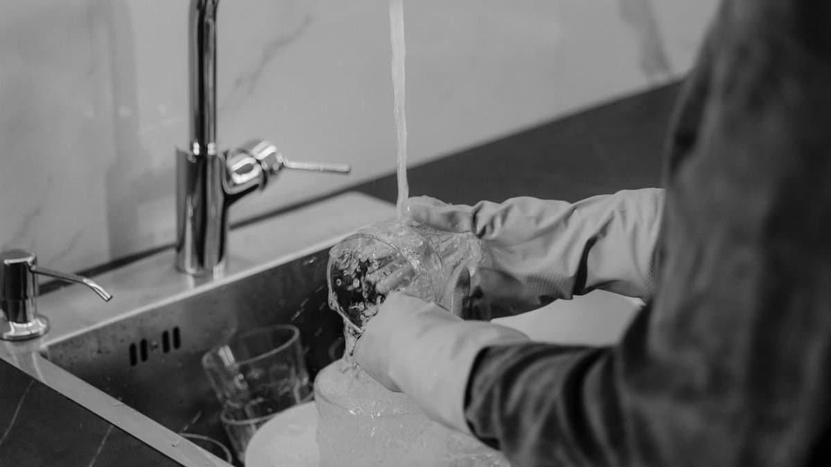 Monochrome photo of a person washing dishes under running water in a kitchen sink.