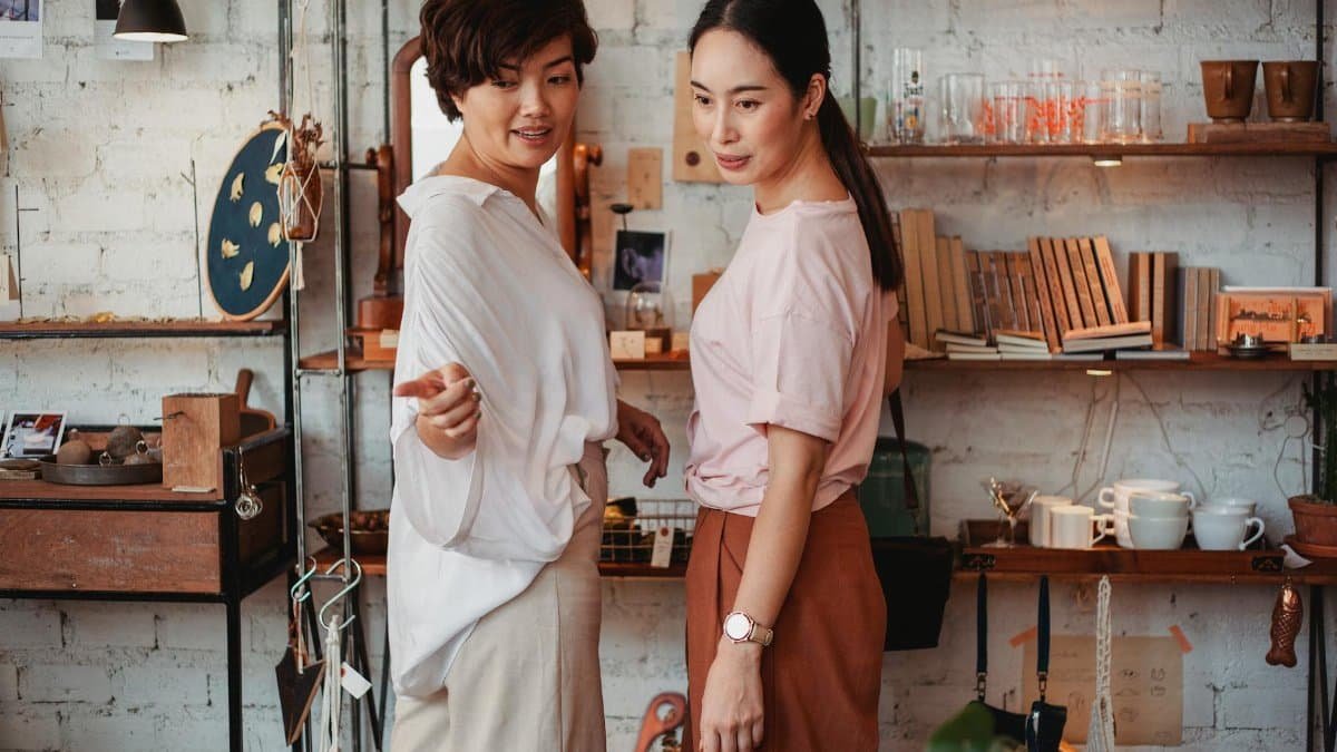 Side view of young Asian buyer pointing with finger near focused girlfriend while selecting kitchen items in store and looking away