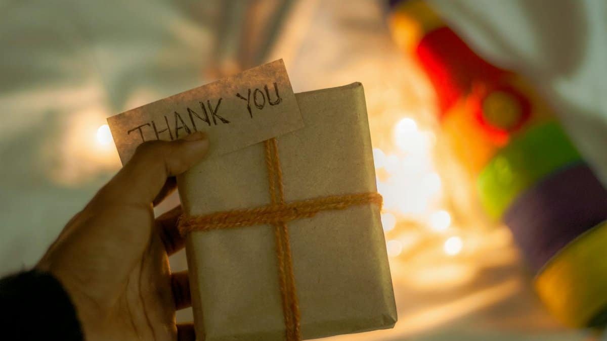 A close-up of a hand holding a gift with a 'Thank You' note, wrapped in brown paper and twine, with warm lights.